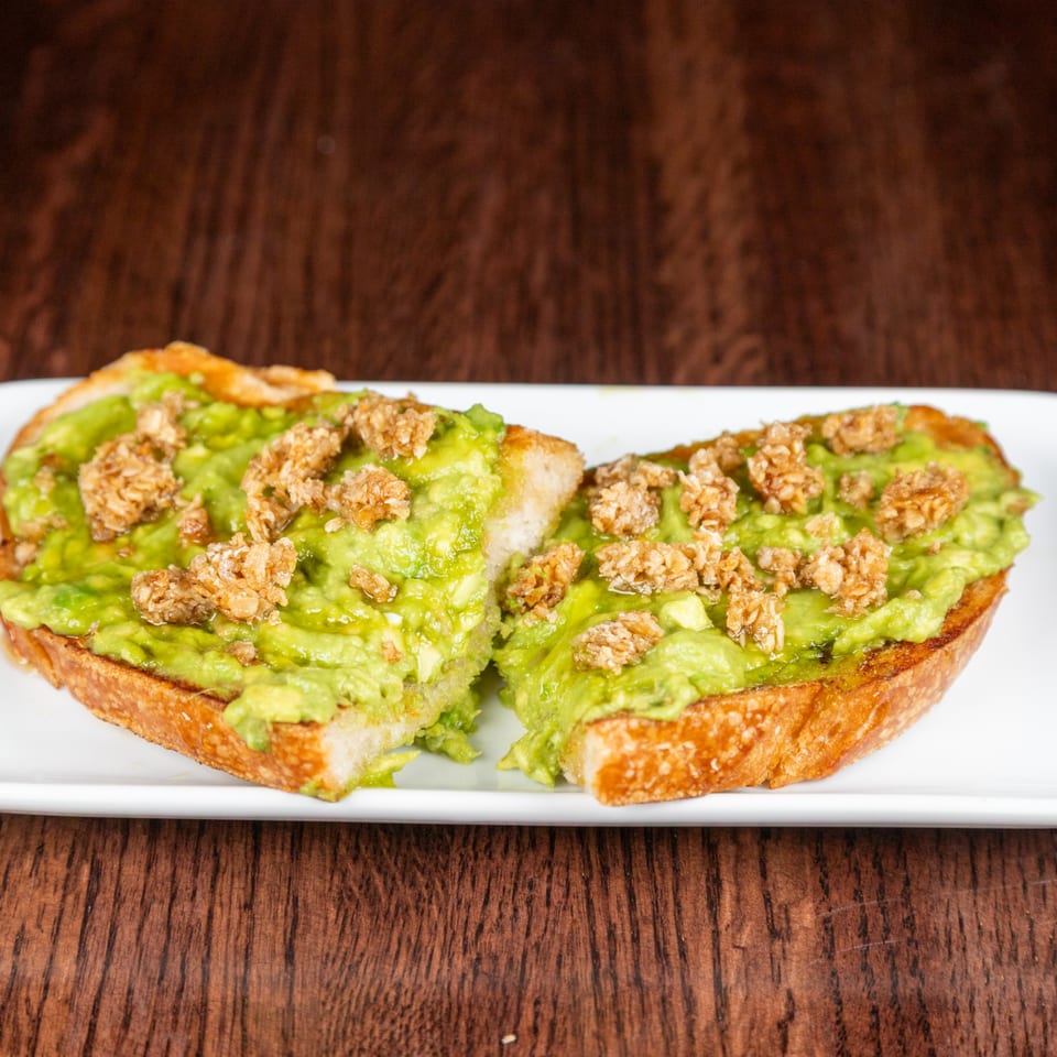 Two slices of avocado toast topped with crunchy granola on a white plate.
