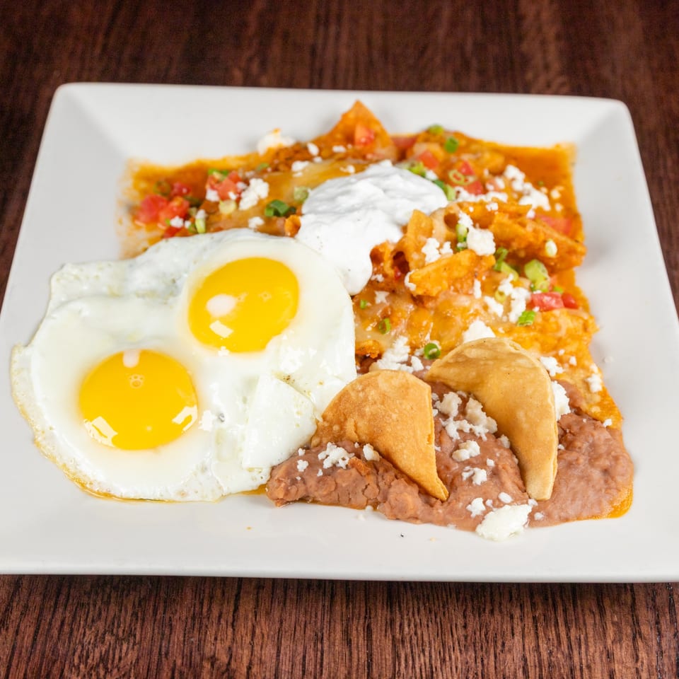 A white plate holds two fried eggs, chilaquiles with sour cream, and refried beans with fried tortillas.
