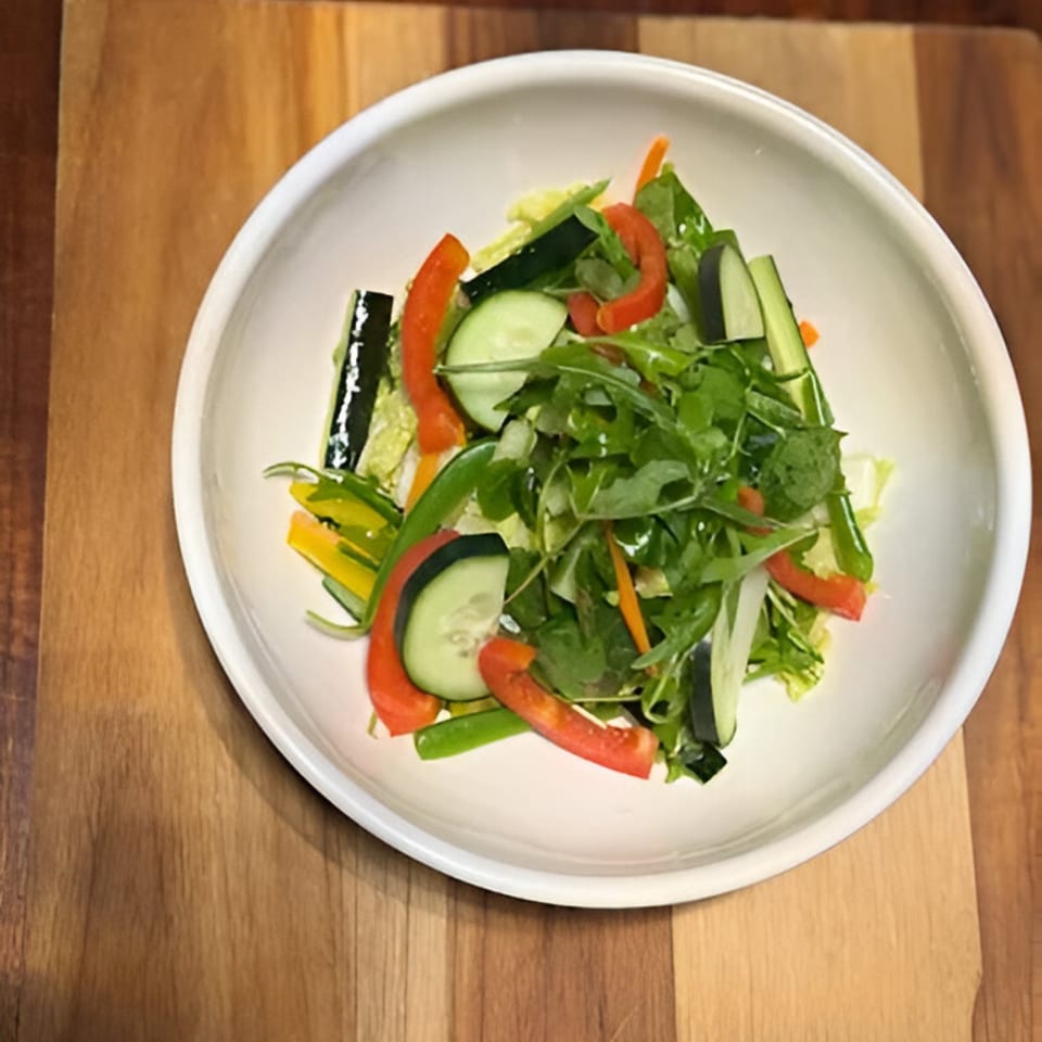 Fresh green salad with cucumbers, red bell peppers, and arugula in a white bowl on a wooden surface.