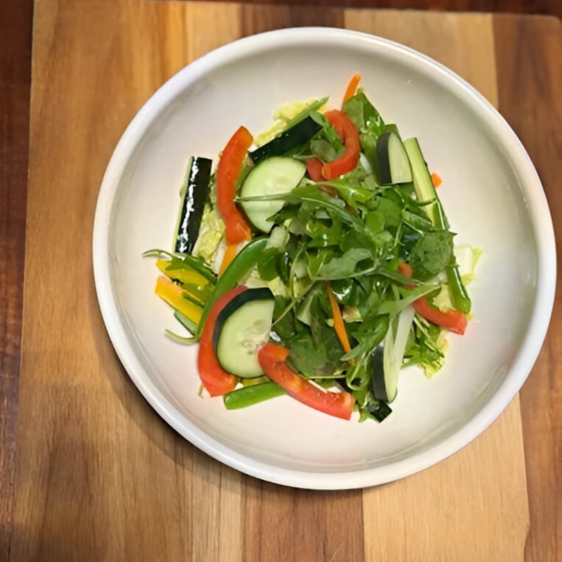 Fresh green salad with cucumbers, red bell peppers, and arugula in a white bowl on a wooden surface.