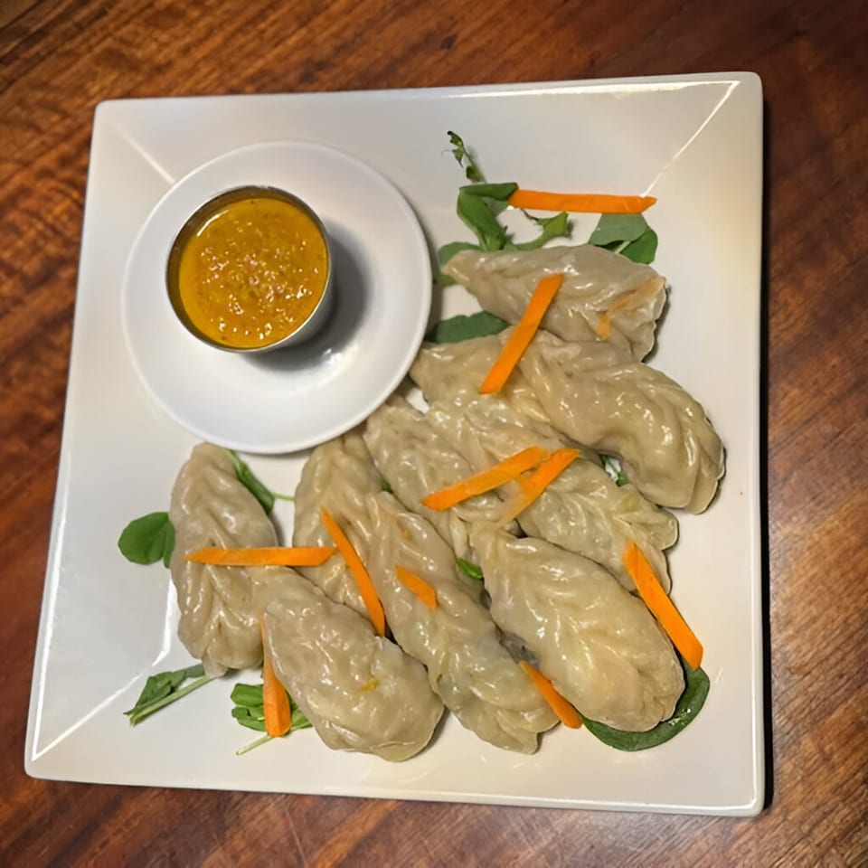 A plate of Nepalese momo dumplings with carrot strips, greens, and a side of orange dipping sauce.