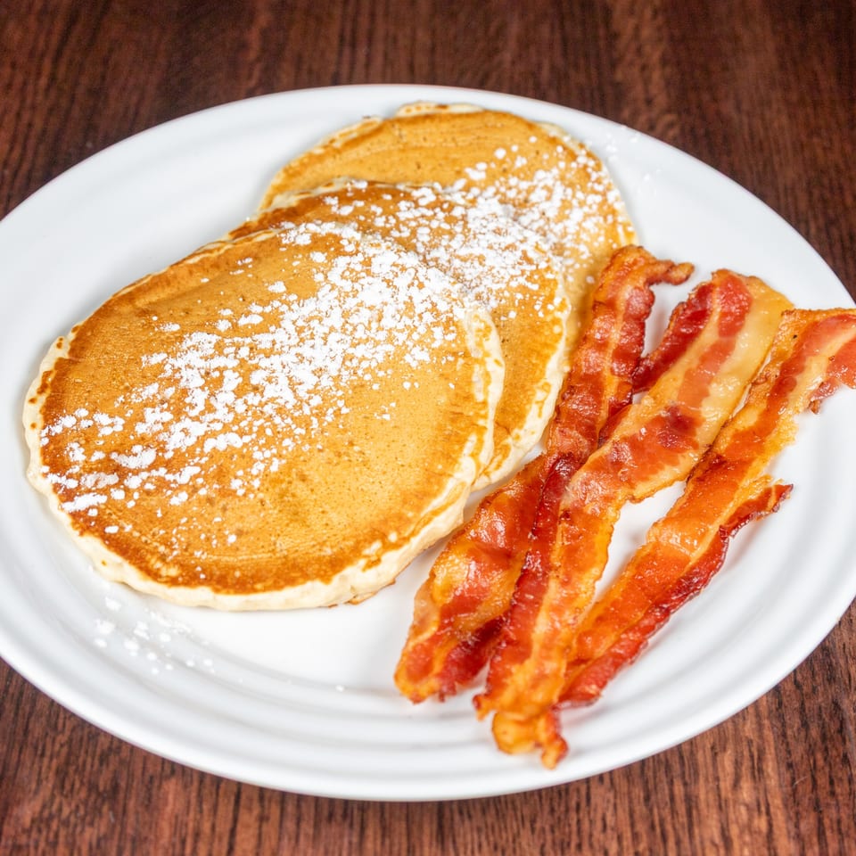 Three golden pancakes dusted with powdered sugar, alongside crispy bacon strips on a white plate.