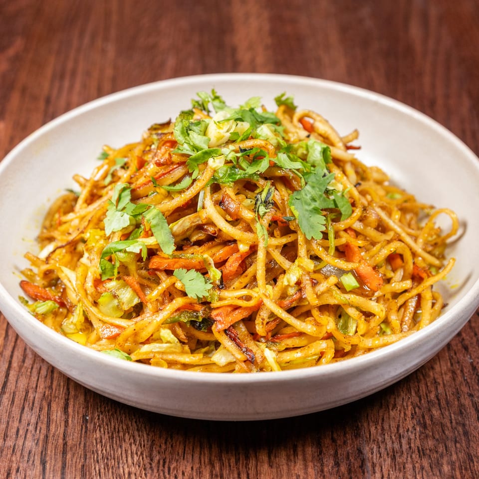 A white bowl of Nepalese chowmein noodles with vegetables and cilantro on a wooden table.