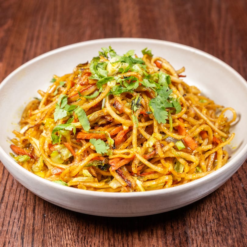 A white bowl of Nepalese chowmein noodles with vegetables and cilantro on a wooden table.